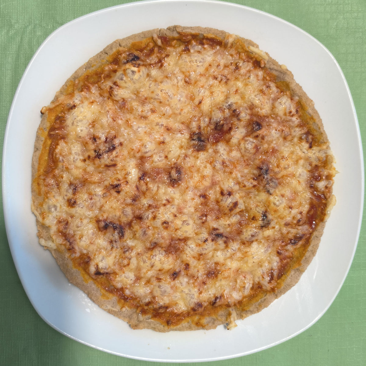 Draufsicht auf eine ganze, rund gebackene Pizza auf einem weißen Teller. Der glutenfreie Boden aus der Bio-Backmischung broat 'das Dünne' ist goldbraun gebacken und mit Tomatensoße und geschmolzenem Käse belegt