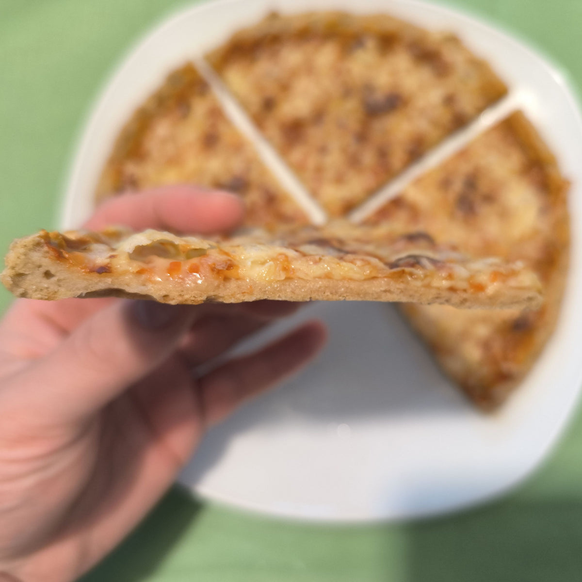 Nahaufnahme eines Pizzastücks der Sorte broat 'das Dünne' in der Seitenansicht. Eine Hand hält das Stück, wodurch der extrem dünne Querschnitt des glutenfreien Bodens sichtbar wird, im Hintergrund die restliche Pizza