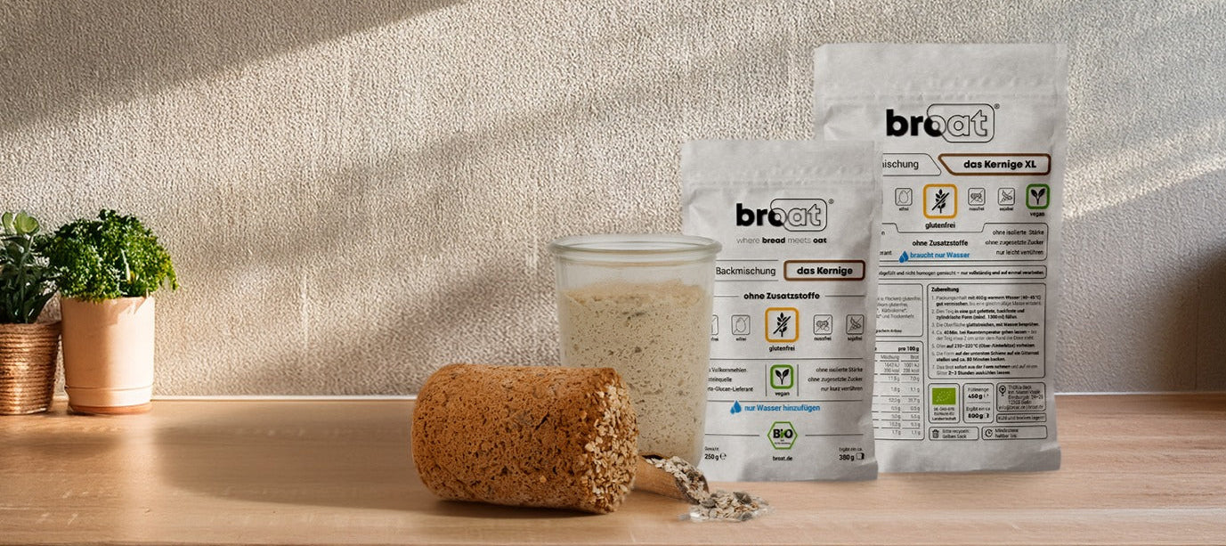 Zwei weiße Verpackungen von broat mit glutenfreien Brotbackmischungen, eine davon die Sorte "das Kernige XL", stehen auf einer Holztheke neben einem Glas mit Backmischungspulver und einem halb angeschnittenen, fertigen, körnigen Brotlaib. Der Hintergrund zeigt eine helle Wand mit sanftem Schatten. Das Bild stammt von broat.de und bewirbt glutenfreie Brotbackmischungen.