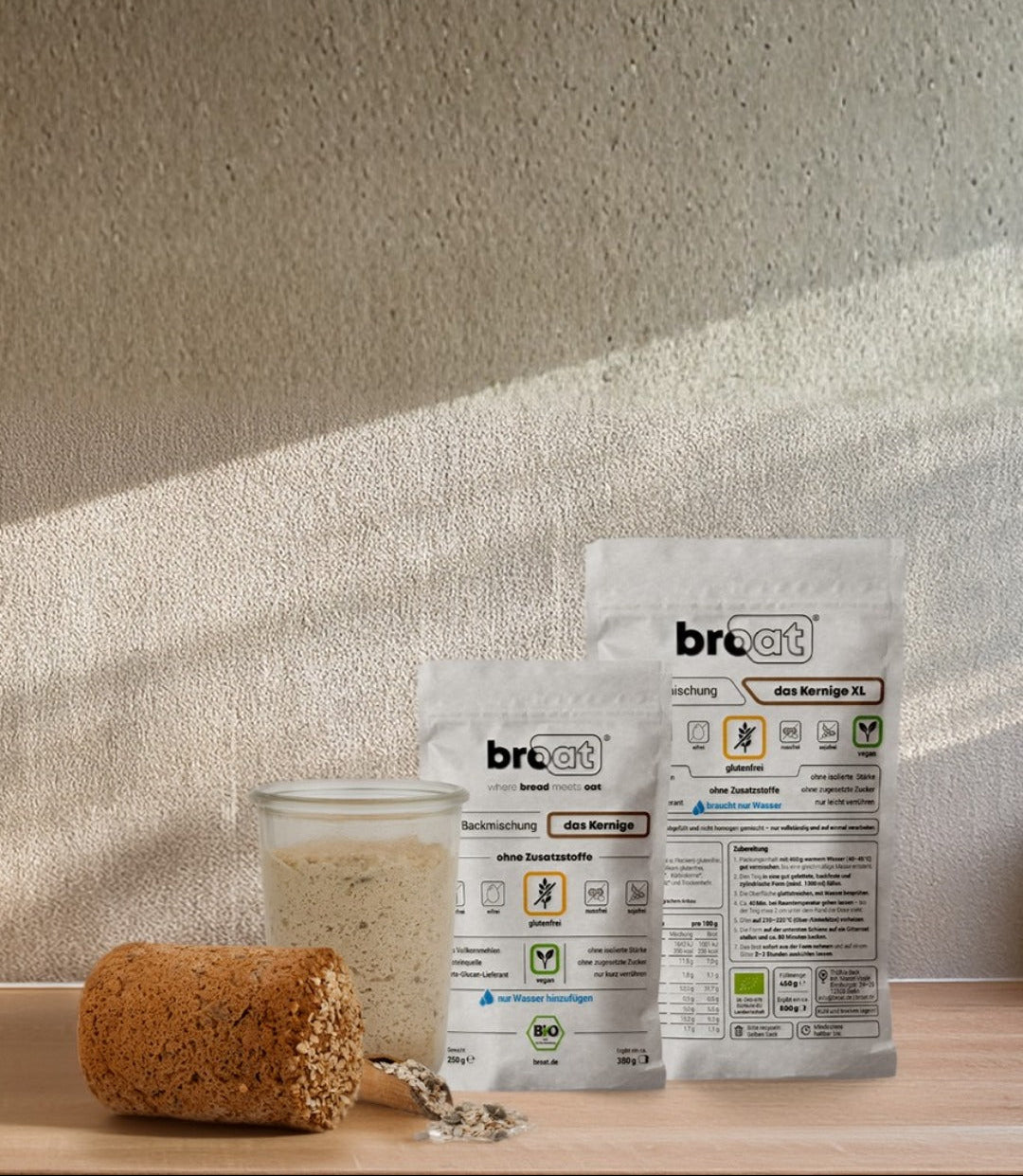 Zwei weiße broat Verpackungen mit glutenfreien Brotbackmischungen, eine davon die Sorte "das Kernige XL", stehen auf einer Theke. Links daneben sind ein Glas mit Teig und ein Anschnitt eines körnigen Brotes zu sehen. Der Hintergrund ist eine helle, strukturierte Wand. Das Bild bewirbt glutenfreie Brotbackmischungen.