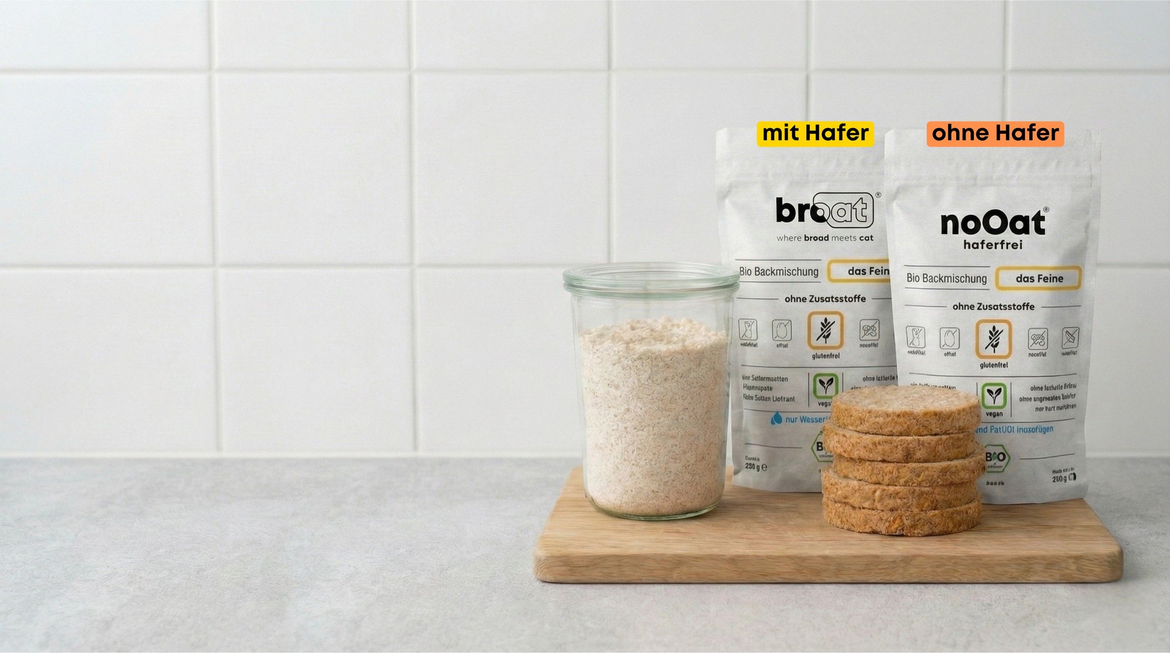 Glutenfreie Bio-Backmischungen der Sorten broat und noOat auf einem Holzbrett, arrangiert neben einem Glas mit der Mischung und drei Scheiben fertig gebackenem Brot ohne Zusatzstoffe