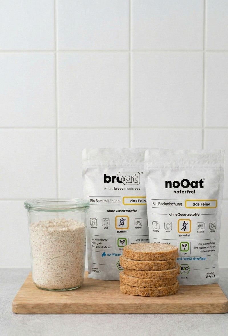 Glutenfreie Bio-Backmischungen der Sorten broat und noOat auf einem Holzbrett, arrangiert neben einem Glas mit der Mischung und drei Scheiben fertig gebackenem Brot ohne Zusatzstoffe